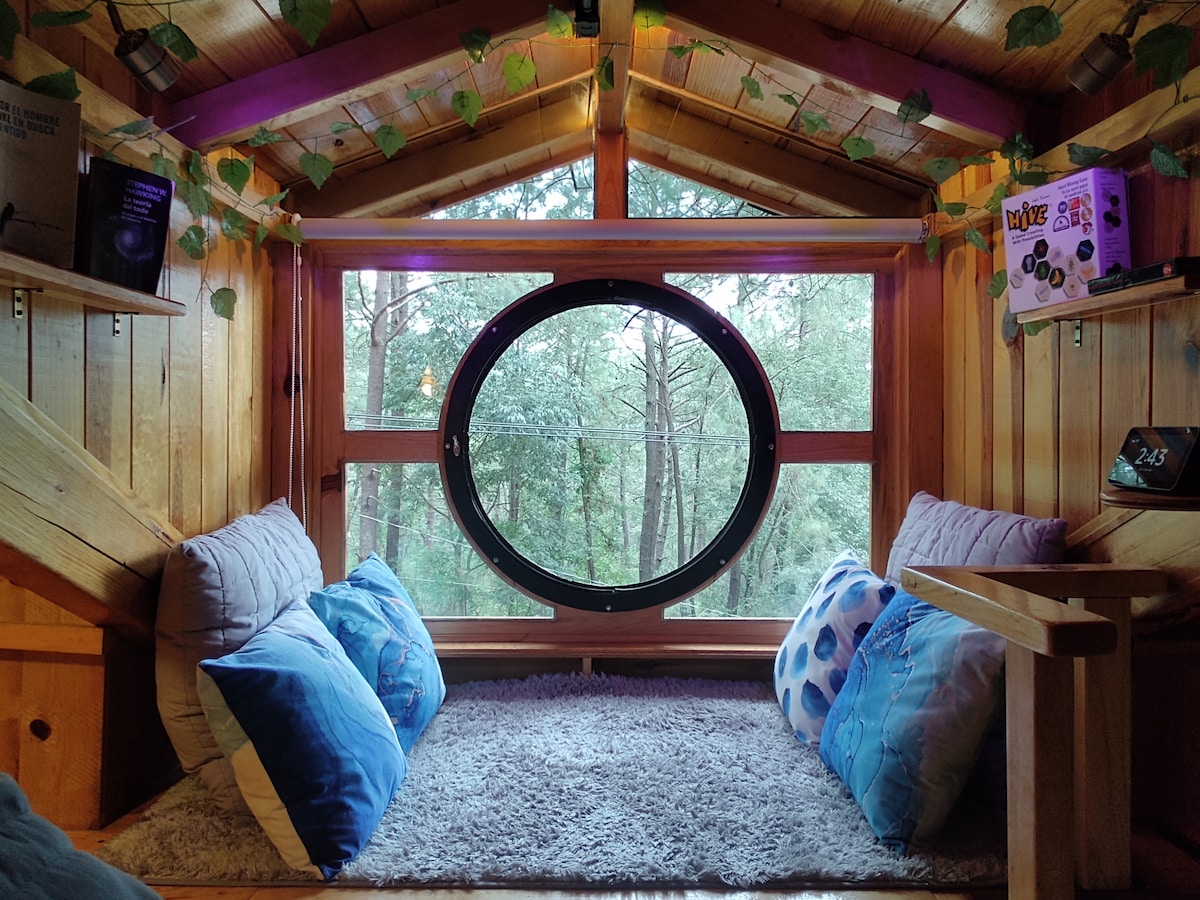A cozy nook is shown with soft cushions placed next to a circular window. Green foliage decorates the wooden walls, adding a natural touch. Plush blue pillows complement the neutral tones of a soft rug, creating a comfortable seating area overlooking the surrounding forest.