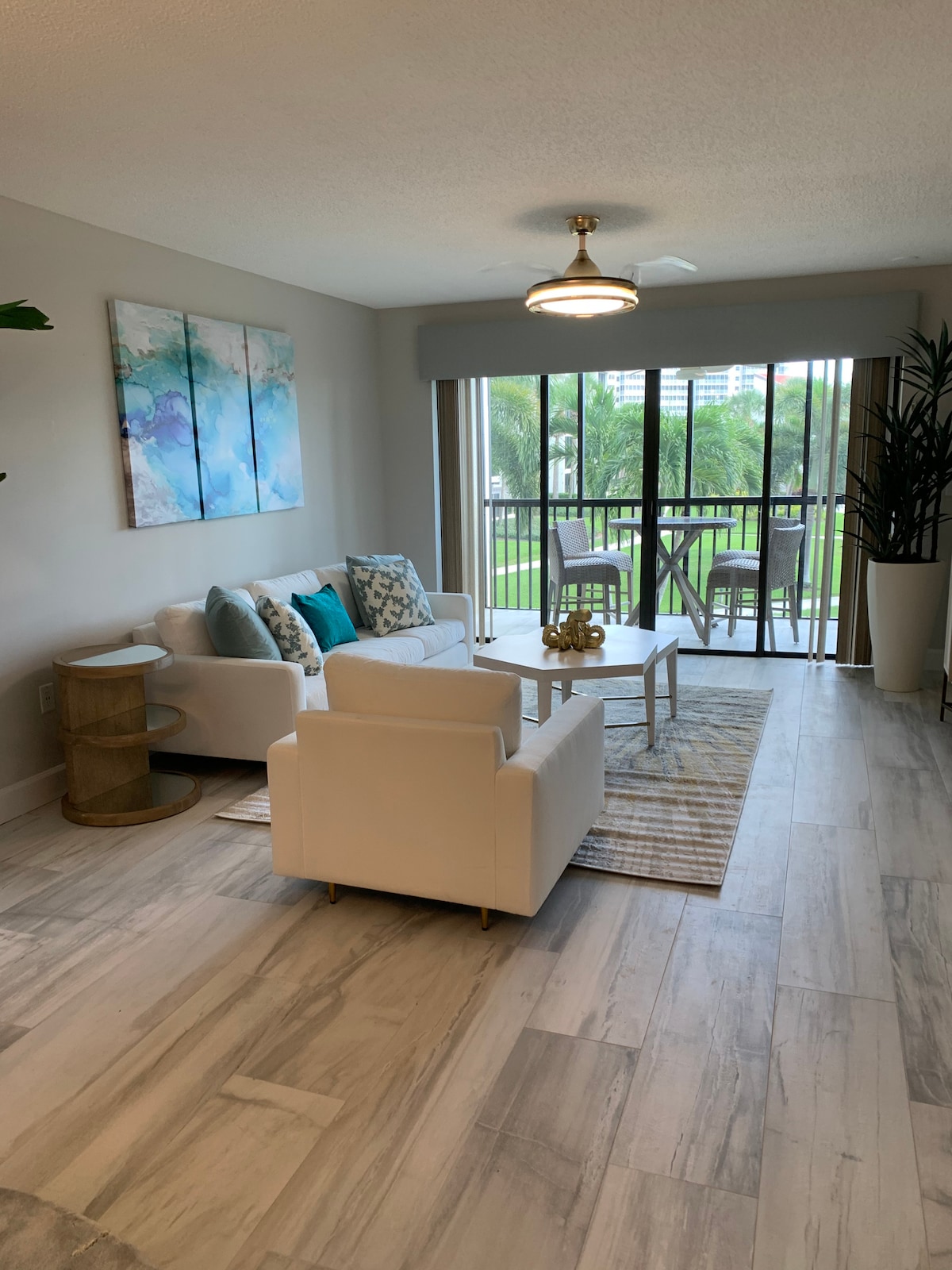 A stylish living area features a light-colored sofa adorned with decorative pillows, accompanied by a matching armchair. A round coffee table is positioned at the center. Natural light enters through large glass doors, which open to a balcony with views of greenery.