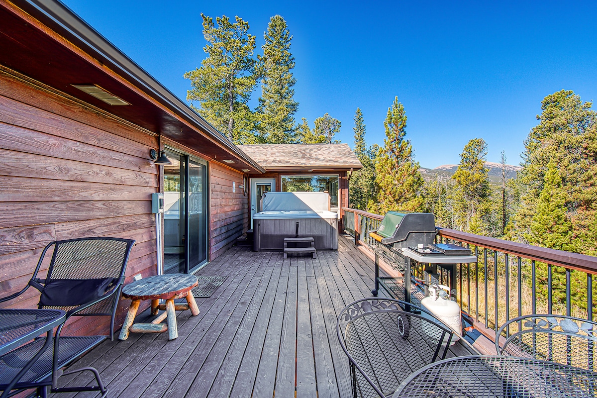 The spacious wooden deck features a hot tub and a grill, surrounded by elevated views of tall trees and mountain peaks. Multiple seating areas include a dining table and lounge chairs, inviting relaxation in a serene outdoor setting.