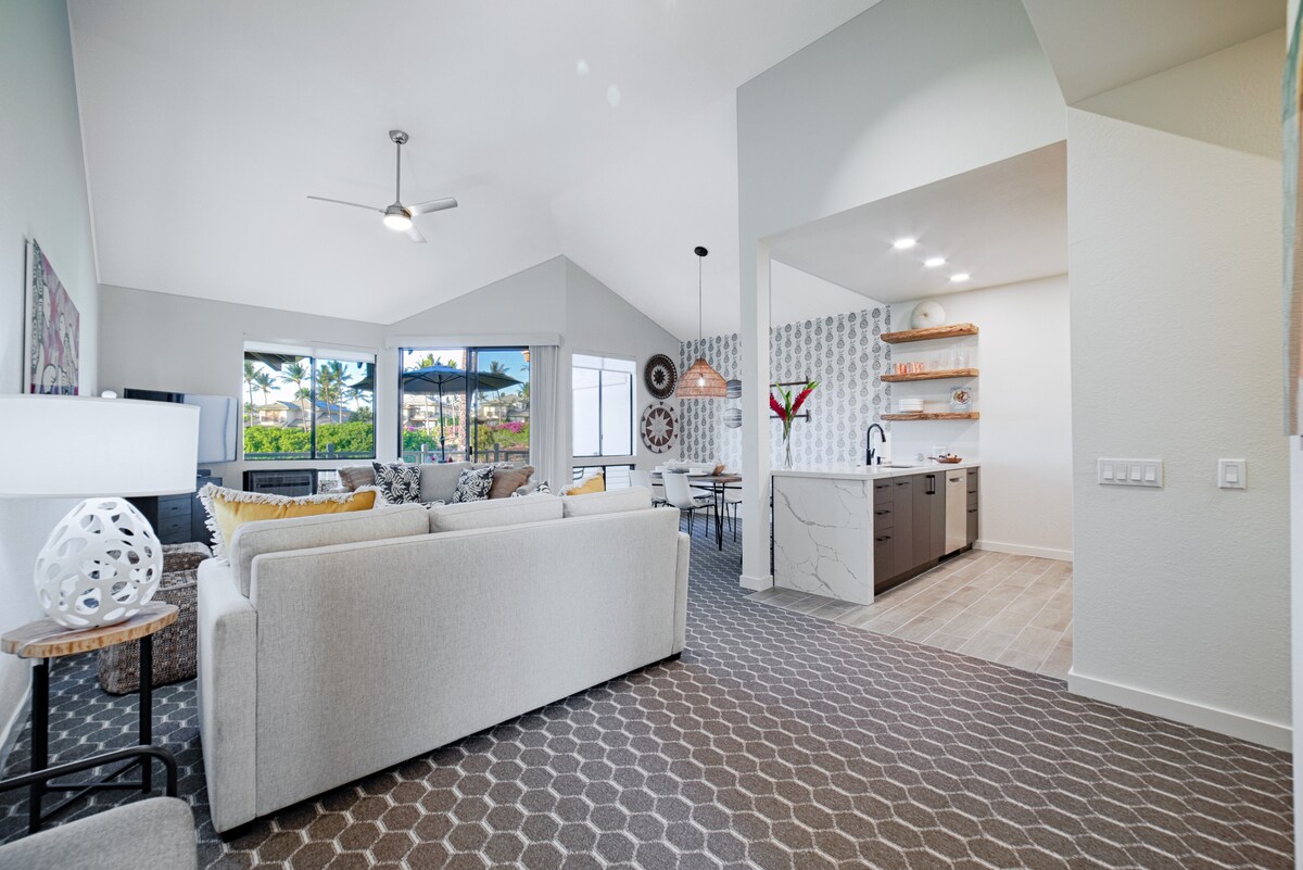 An inviting living area showcases a spacious couch with colorful cushions, positioned near large windows that frame views of the outdoors. The adjacent kitchen features sleek countertops and modern shelving, enhancing the contemporary aesthetic of the open floor plan.