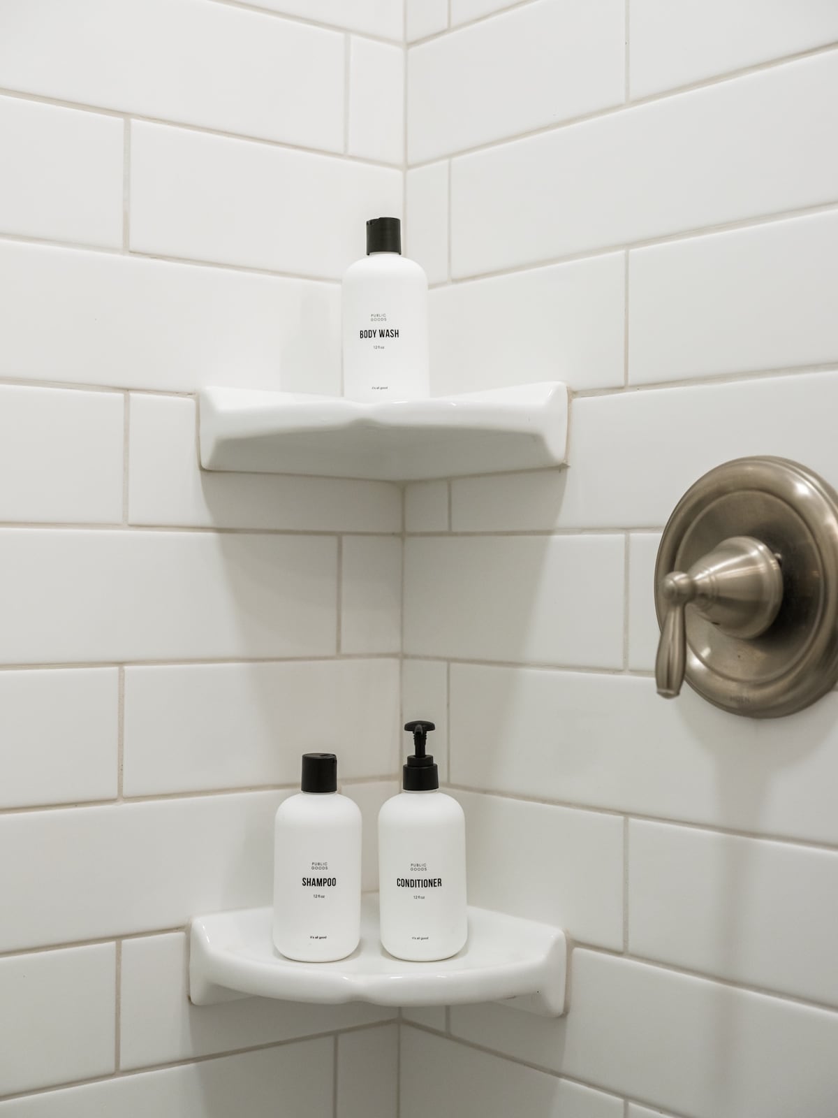 A clean and modern shower features two white shelving units displaying body wash, shampoo, and conditioner in simple, minimalist bottles. The white tiled walls provide a fresh backdrop, while a polished metal shower faucet adds a touch of elegance.
