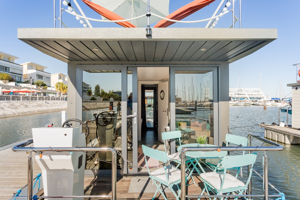 A contemporary houseboat is shown from the exterior, featuring large glass doors leading into the living space. A seating area with light blue chairs is arranged on the deck, alongside a helm station. Boats and marina scenery can be seen in the background.