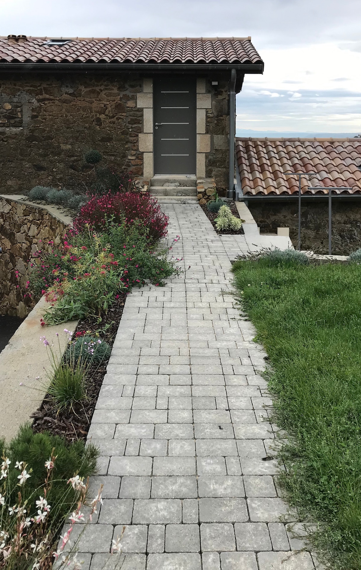 A stone pathway is lined with colorful flowers and greenery, leading to a neutral-toned entrance door. The rustic texture of the stone walls and terracotta roof tiles is complemented by a well-maintained grassy area on one side.