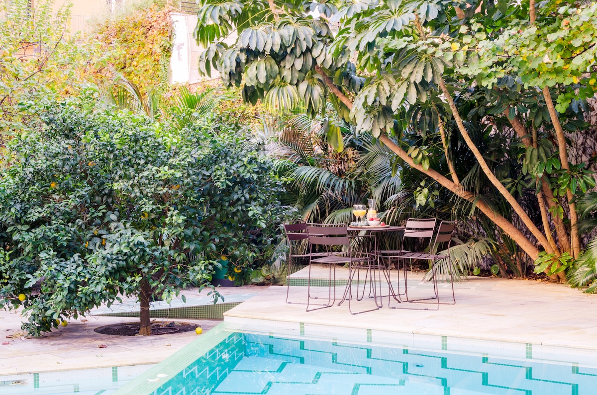 A serene outdoor area features a sparkling pool surrounded by lush greenery. A small table with four chairs is positioned nearby, offering a space for relaxation. The area is enriched by tropical plants, creating a tranquil setting for guests to enjoy.