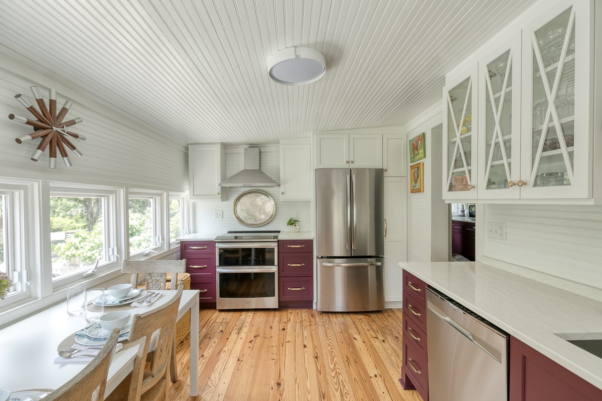 The kitchen features modern stainless appliances, including a refrigerator and range. White cabinetry contrasts with deep purple accents. A dining table set for four is positioned by large windows, allowing natural light to stream in, while polished wooden floors add warmth to the space.