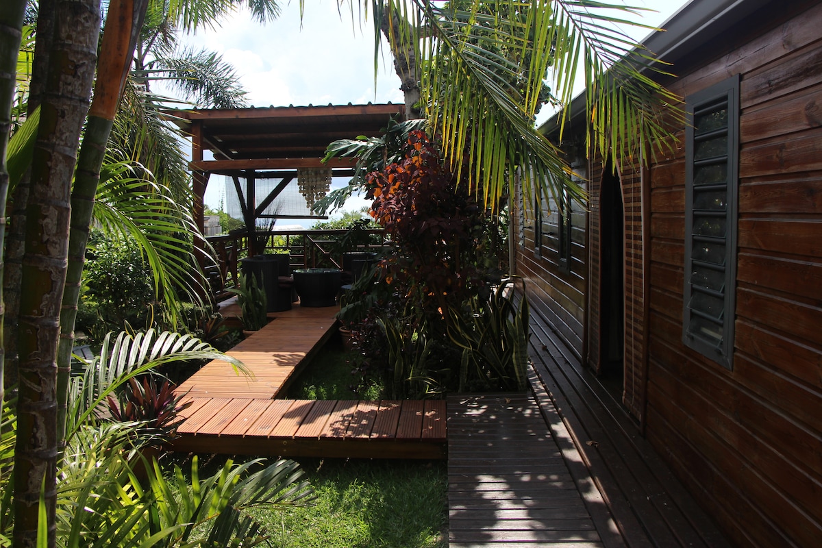 A wooden walkway meanders through lush greenery, leading to a charming cabin. Tropical plants frame the pathway, while a terrace offers views of the surrounding garden. Sunlight filters through the leaves, creating a serene atmosphere in this private outdoor space.