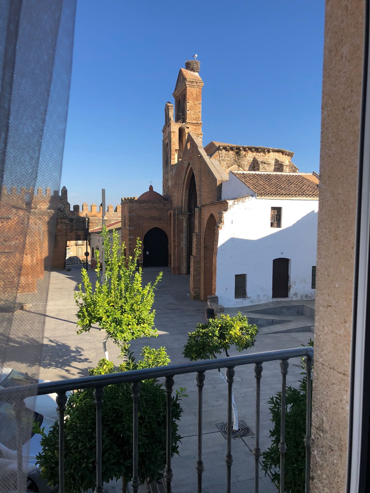 The image captures a view from a balcony overlooking a charming historic square. A brick church with a tall bell tower stands prominently in the background, while a small white building is visible nearby. Lush green trees line the square, enhancing the scene's vibrancy.