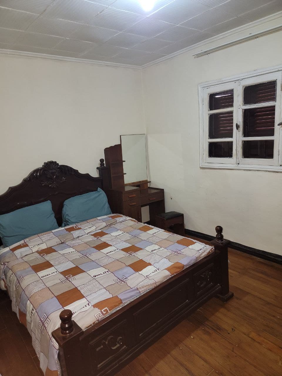 A simple bedroom is furnished with a spacious bed covered in a patchwork blanket. Light-colored walls enhance the room's brightness, and a window with wooden shutters allows natural light to filter in. A dresser is positioned beside the bed, providing additional storage.