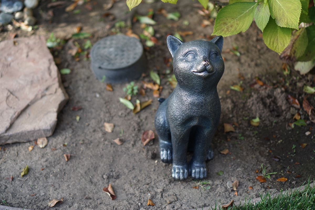 A whimsical garden sculpture of a cat sits amidst fallen leaves and dirt, surrounded by greenery. The cat's cheerful expression and textured surface invite a sense of playfulness to the outdoor space.