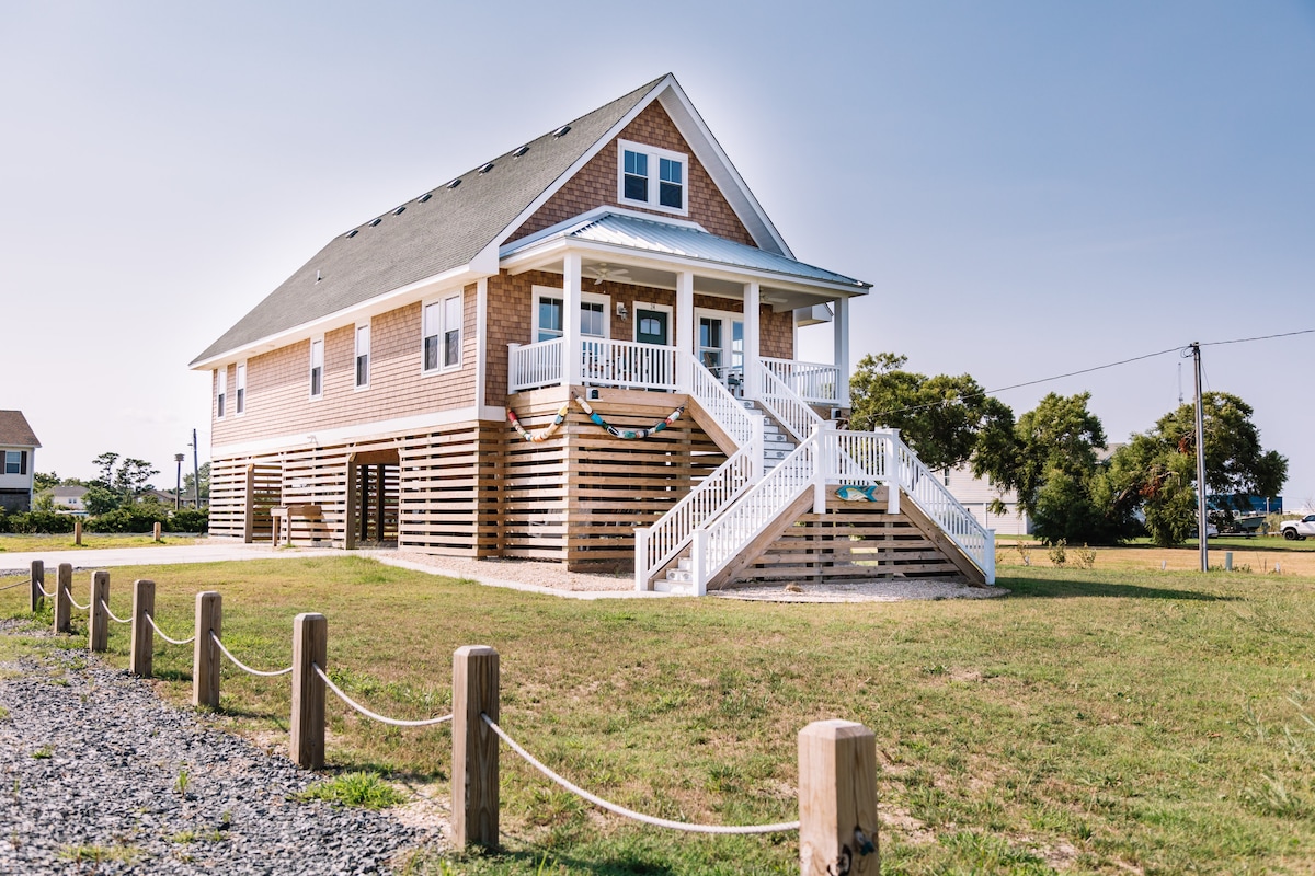 The cottage features a light wood exterior with a welcoming front porch and white railings. Stairs lead up to the entrance, while the ground level is supported by stilts. The surrounding grassy area provides space for outdoor activities and is lined with wooden posts.