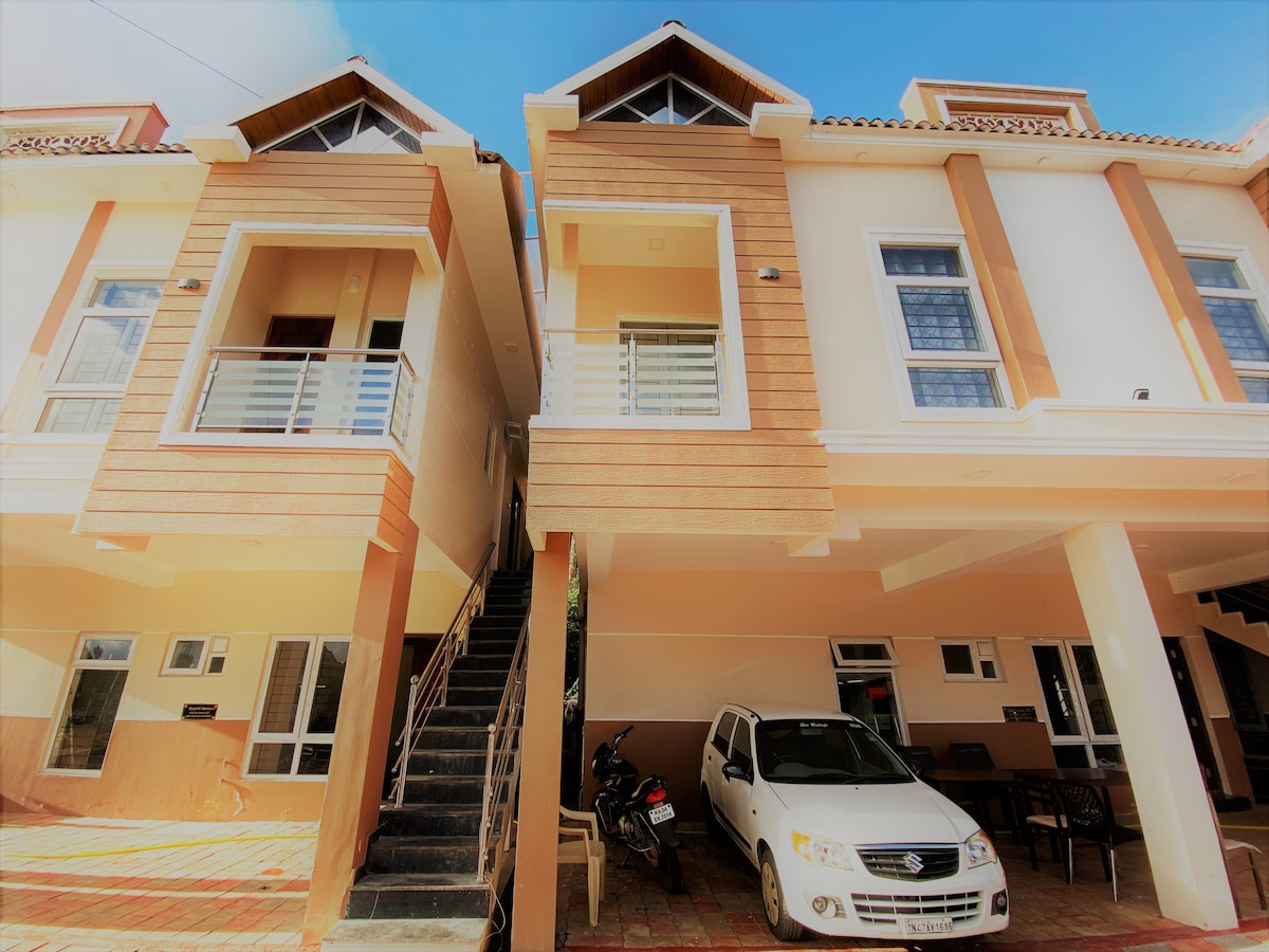 The exterior features a pair of townhouses with wooden accents. A staircase leads to the upper level. Directly below, a parking area accommodates a vehicle and a motorcycle, while outdoor seating is visible on a paved surface.