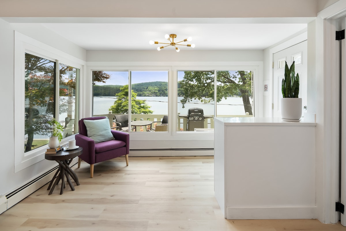 A cozy sitting area features a purple armchair with a soft green cushion, positioned by large windows that offer views of the serene lake. Natural light fills the space, highlighting the light wooden flooring and modern light fixture above.