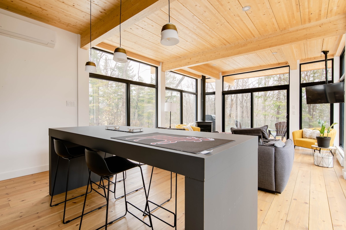 A modern dining area features a large, minimalistic table surrounded by sleek black chairs. Natural light floods in through floor-to-ceiling windows, illuminating wooden ceiling beams and the warm-toned hardwood floor. A cozy lounge area with comfortable seating is visible in the background.