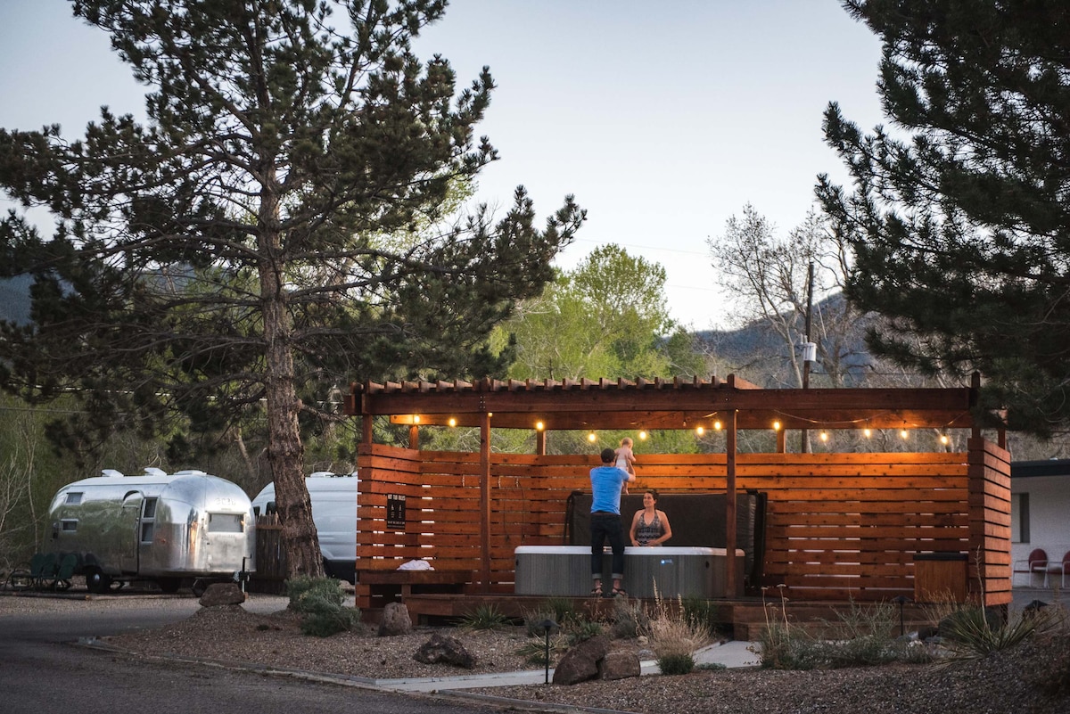 An outdoor hot tub is situated beneath a wooden gazebo, enhanced by warm string lights. A couple enjoys the space, surrounded by lush greenery and tall trees. In the background, classic silver Airstream trailers are visible against a serene landscape.