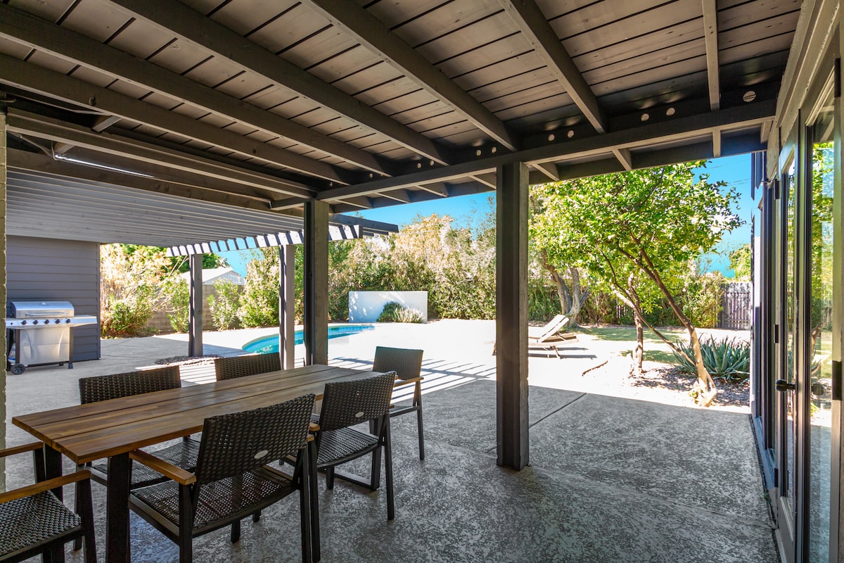 A covered patio area features a dining table surrounded by chairs, offering a shaded space for relaxation. In the background, a private heated pool is visible, alongside lounge chairs and a well-maintained grass area, complemented by green landscaping.