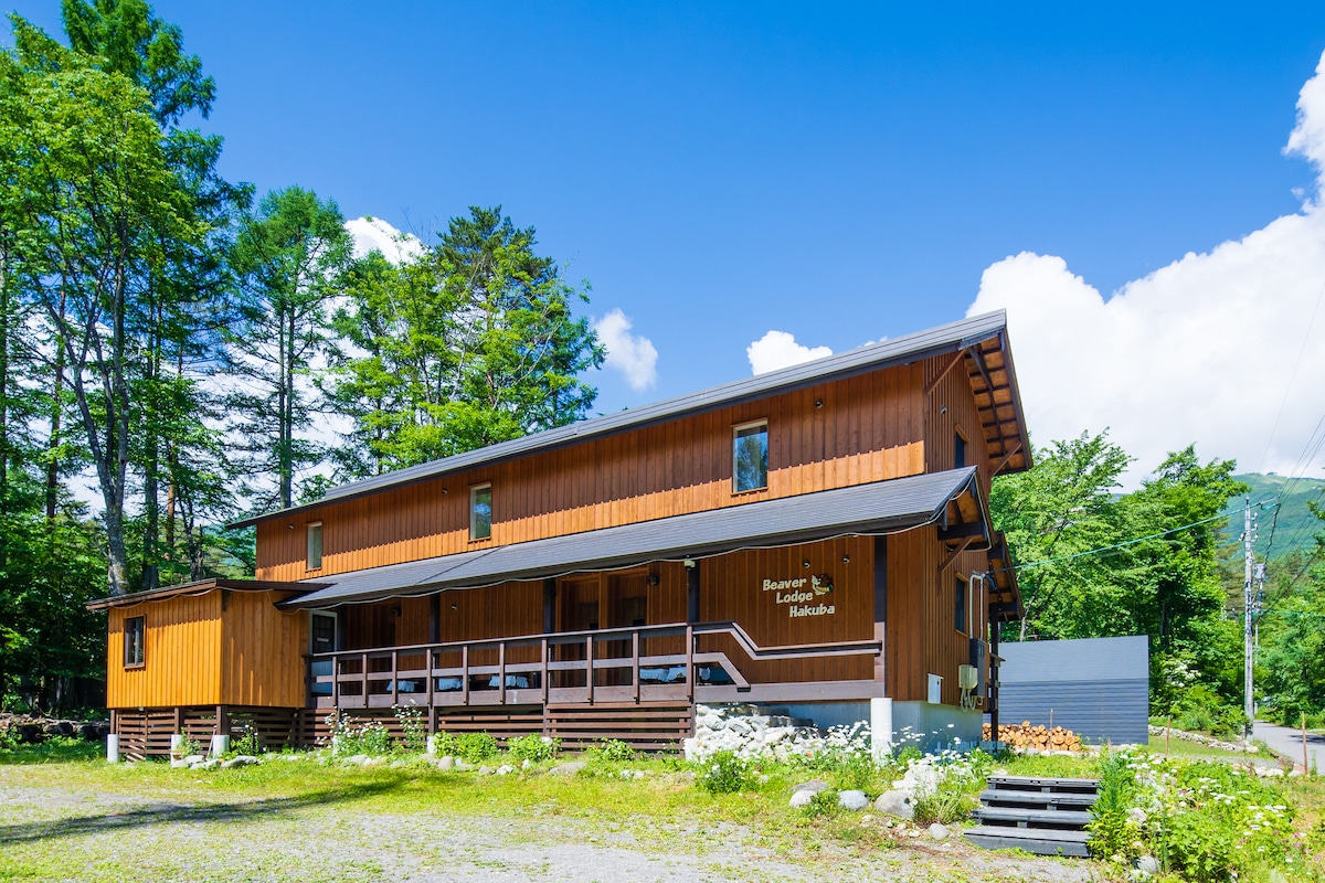 A three-unit chalet is set amidst lush greenery, featuring a wooden facade and expansive front porch. Large windows allow ample natural light, enhancing the connection to the surrounding nature. The structure's two stories offer a spacious appearance against a backdrop of a clear blue sky.
