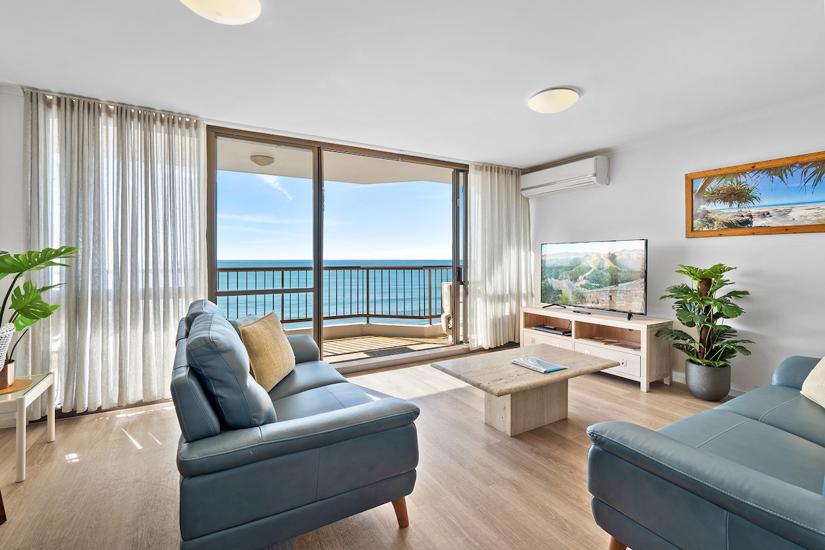 A comfortable living area features two blue sofas positioned towards a flat-screen TV. Sunlight filters through sheer curtains, illuminating the space and revealing stunning ocean views from the large glass balcony doors. A small, neutral-toned coffee table complements the modern decor.