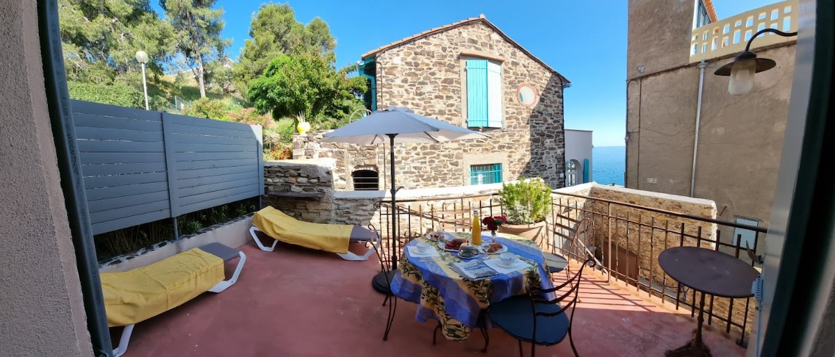 A private patio features a stone wall background and light-colored outdoor furniture. A round dining table is set with dishes, surrounded by chairs. An umbrella provides shade, while two loungers are positioned for relaxation. The area offers views of the surrounding greenery and blue sky.
