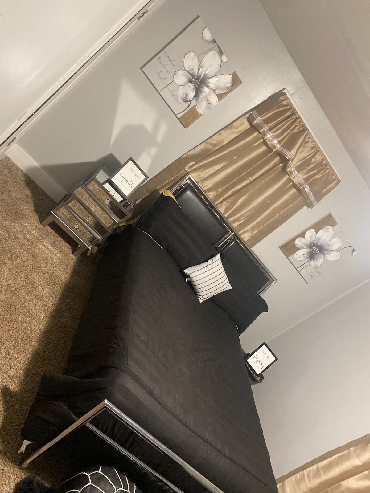 A bedroom features a spacious bed with a black coverlet and decorative throw pillow. Nearby, a nightstand holds framed artwork and a small lamp. Light beige curtains complement the floral wall art above the bed, while a plush brown carpet adds comfort to the space.