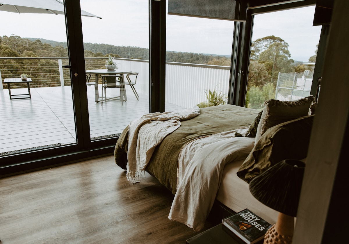 A spacious bedroom features a comfortable bed dressed in neutral tones. Large glass doors open onto a deck, revealing scenic views of the surrounding nature. A small dining table is visible outside, with a lamp and books present on a nearby bedside table.