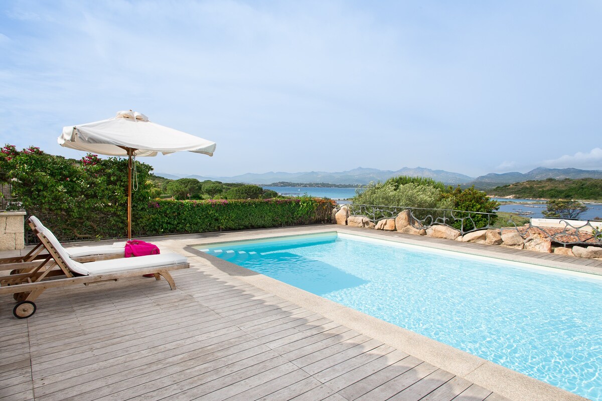 A private pool is surrounded by a wooden deck, with a single sun lounger and an umbrella positioned nearby. Greenery lines the perimeter, complementing the scenic view of the ocean and mountains in the background.