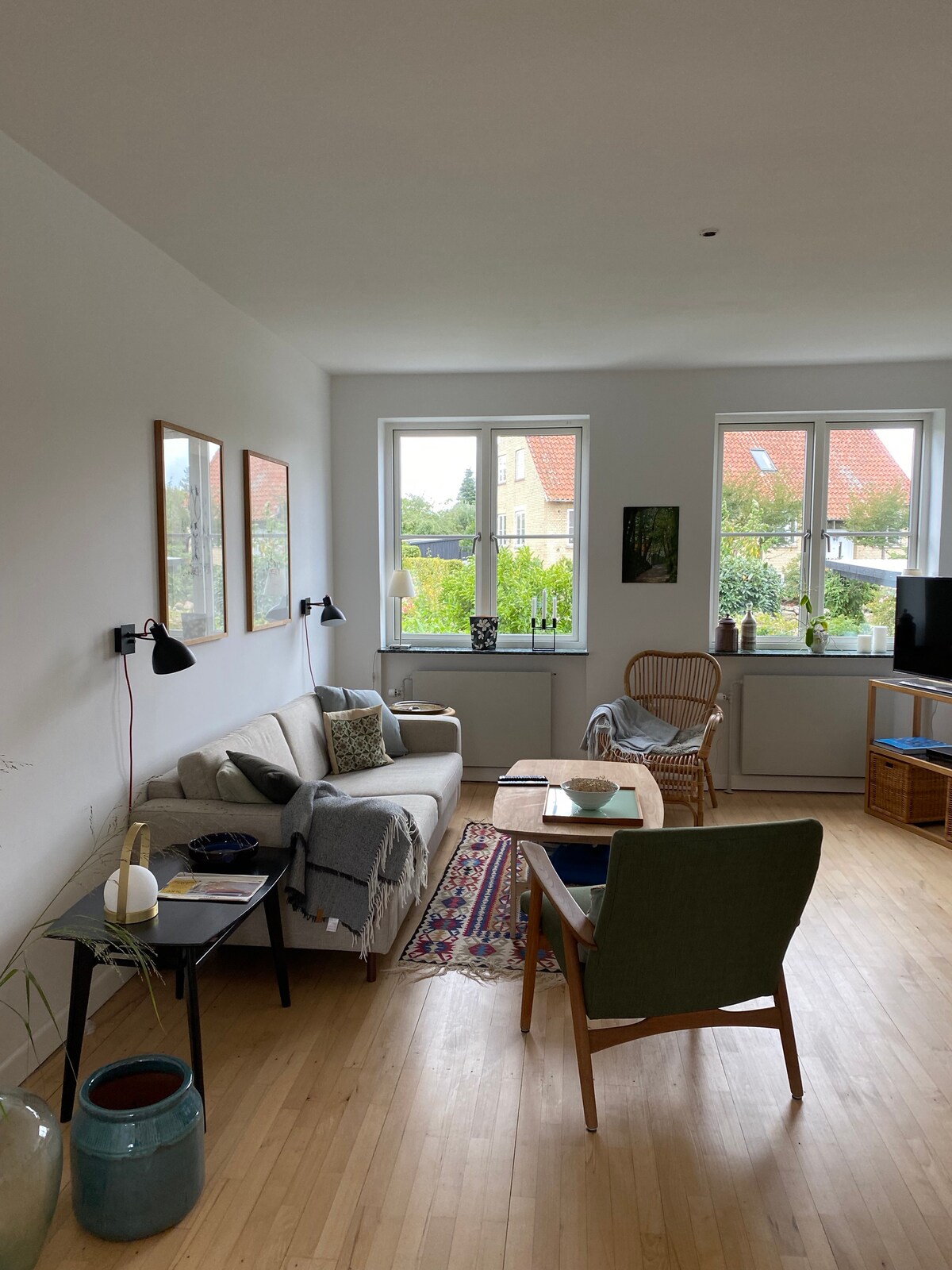 A well-designed living area features a light-colored sofa accompanied by a cozy throw. Two large windows allow natural light to brighten the space, while a small table is positioned between a modern chair and a decorative plant. Artwork is displayed on the walls.