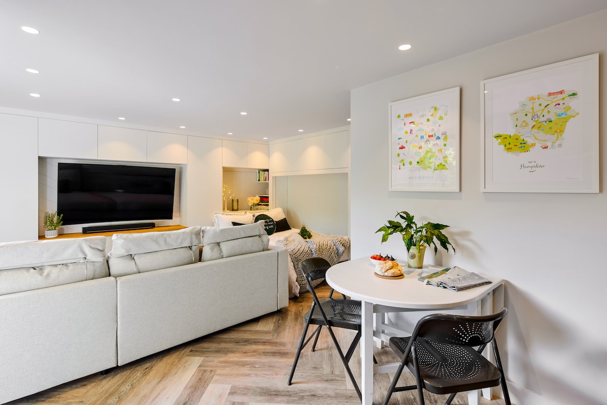 An open living area features a white sectional sofa facing a large flat-screen TV. A dining table with black chairs sits nearby, accompanied by decorative maps and plants on the walls, providing a modern and functional space for relaxation and dining.