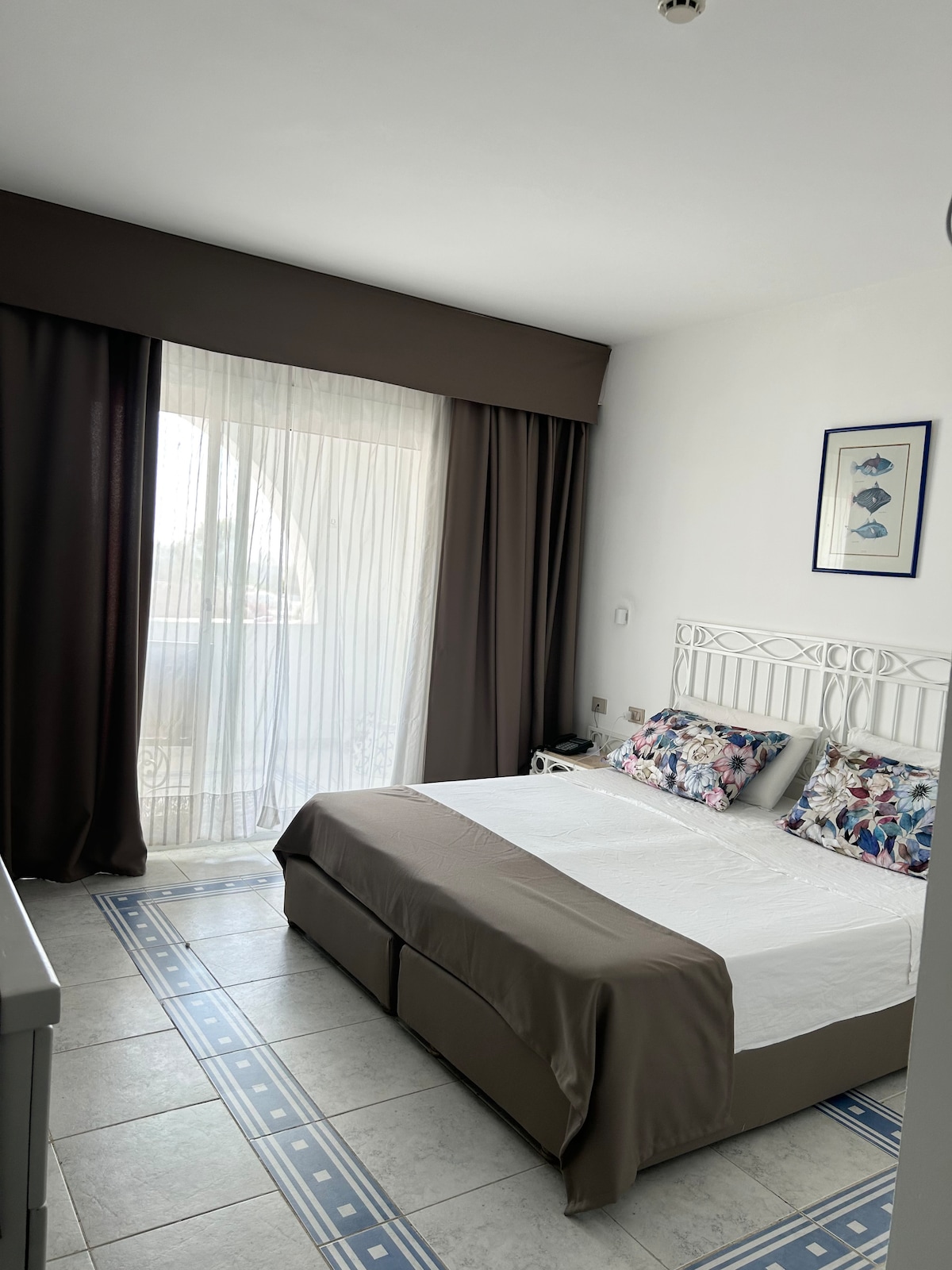 The sleeping area features a spacious bed with a neutral-colored coverlet and floral accent pillows. Natural light filters through sheer curtains, creating a soft glow. A balcony door provides access to the outdoor space, enhancing the room's airy feel.