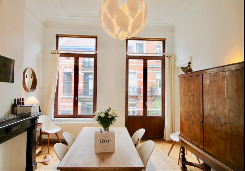 A bright dining area features a large wooden table surrounded by six chairs. Two tall windows provide natural light, and a decorative light fixture hangs from the ceiling. A floral centerpiece is placed on the table, and a wooden cabinet is situated against the wall.
