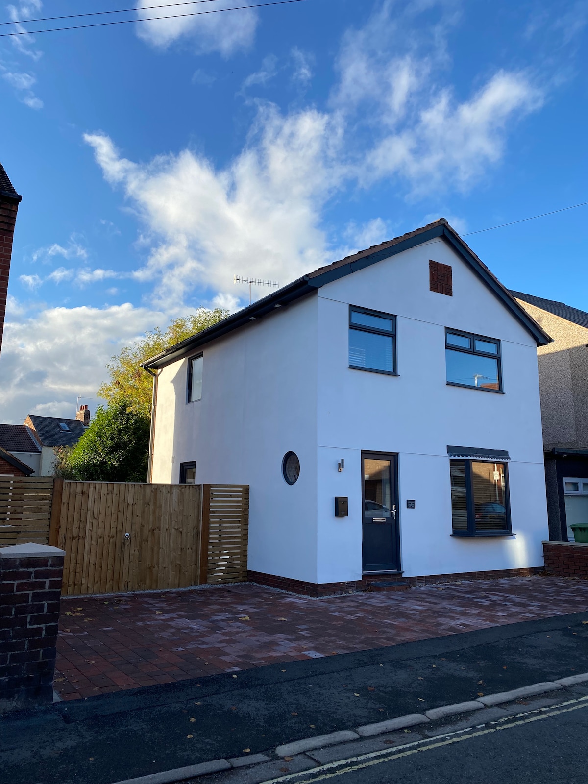 A modern two-story home features a clean white exterior with large windows and a circular accent window. A wooden fence offers privacy to the front yard, which displays a paved pathway leading to the entrance. The sky is partly cloudy, creating a serene backdrop.