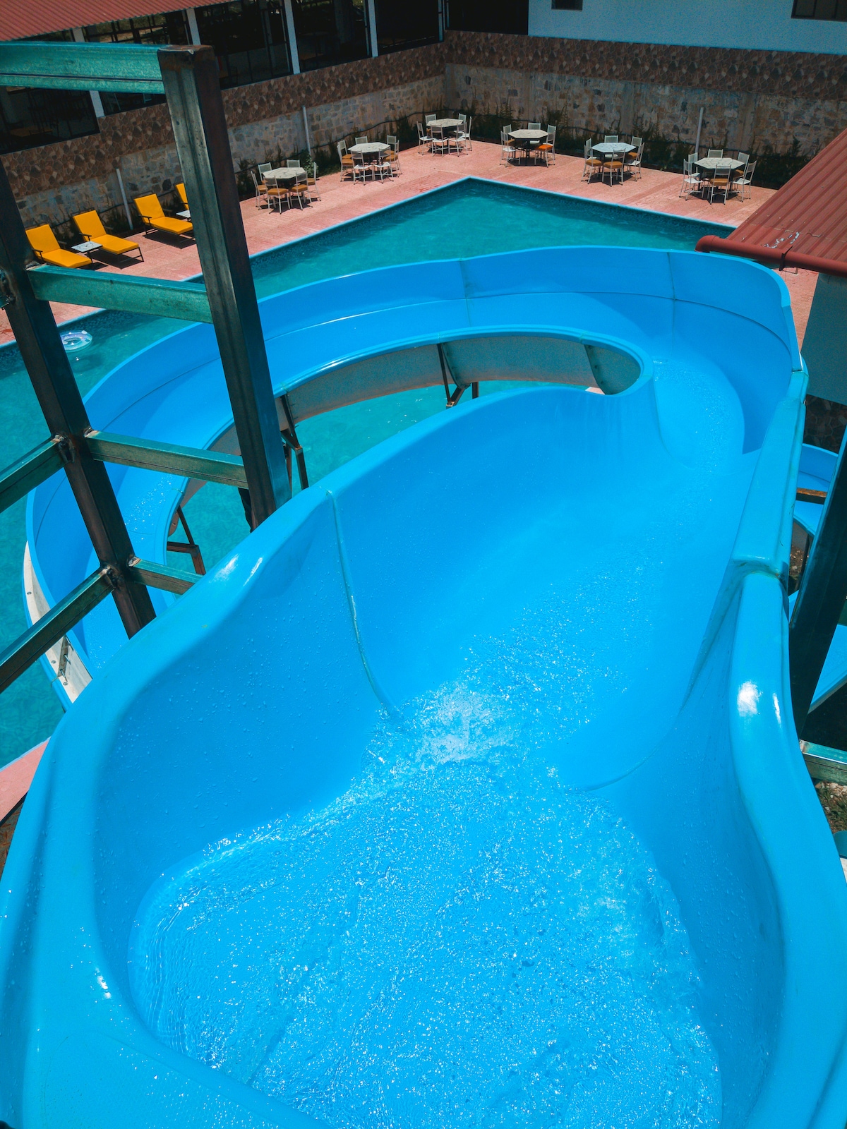 A vibrant blue waterslide descends into a sparkling pool area, framed by sun lounges and shaded tables. The clear water reflects sunlight, creating a refreshing atmosphere for relaxation and fun.