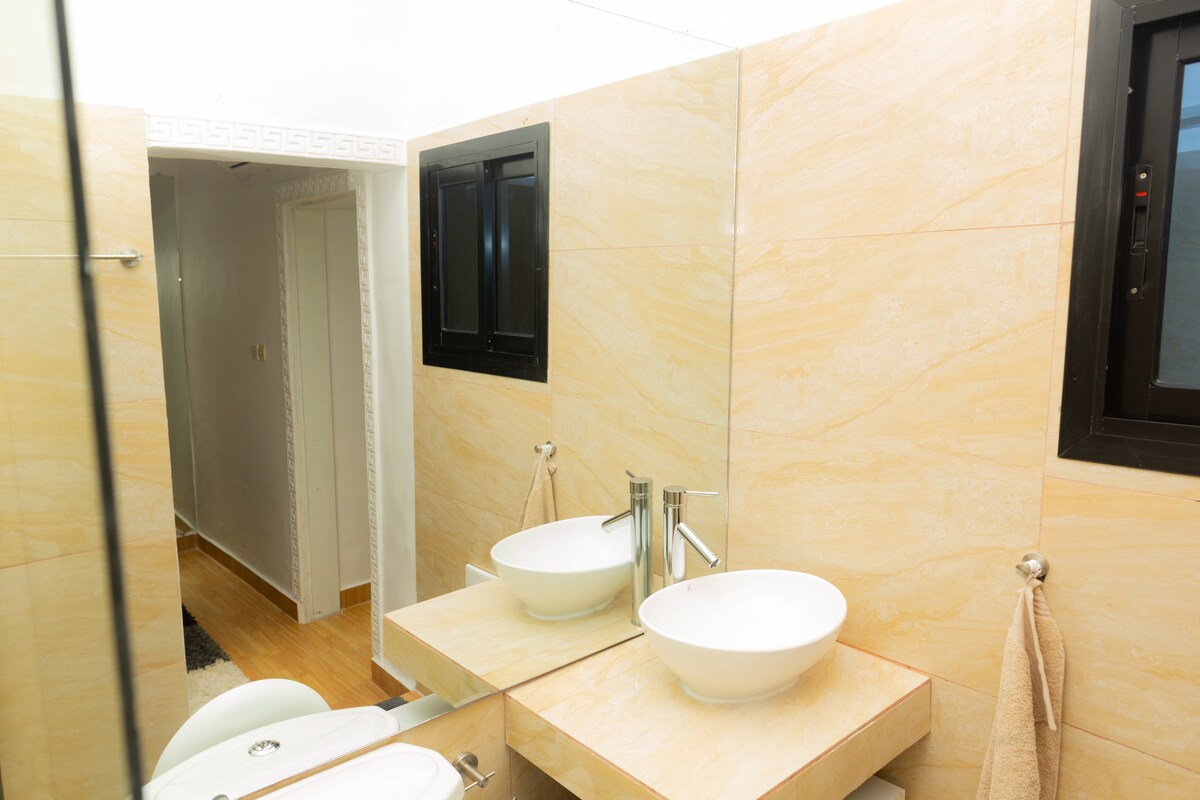 The bathroom features a sleek design with light-colored stone walls. Two modern vessel sinks are mounted on a vanity, complemented by elegant fixtures. Natural light enters through a window set above the sinks, and a towel is neatly hung nearby.