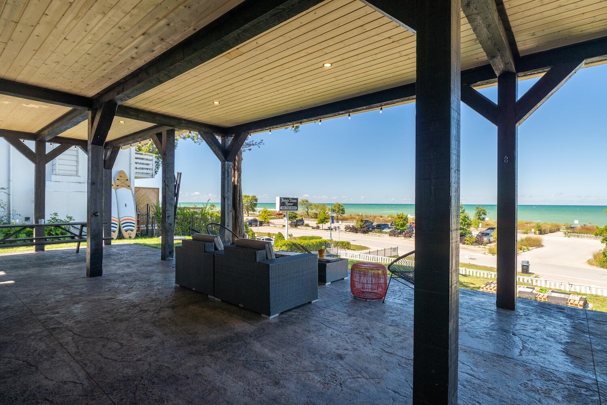 A spacious covered patio provides a relaxing seating area with comfortable furniture, overlooking the nearby beach and water. Clear views of the ocean are visible through the pillars, creating an inviting outdoor space for leisure and enjoyment.