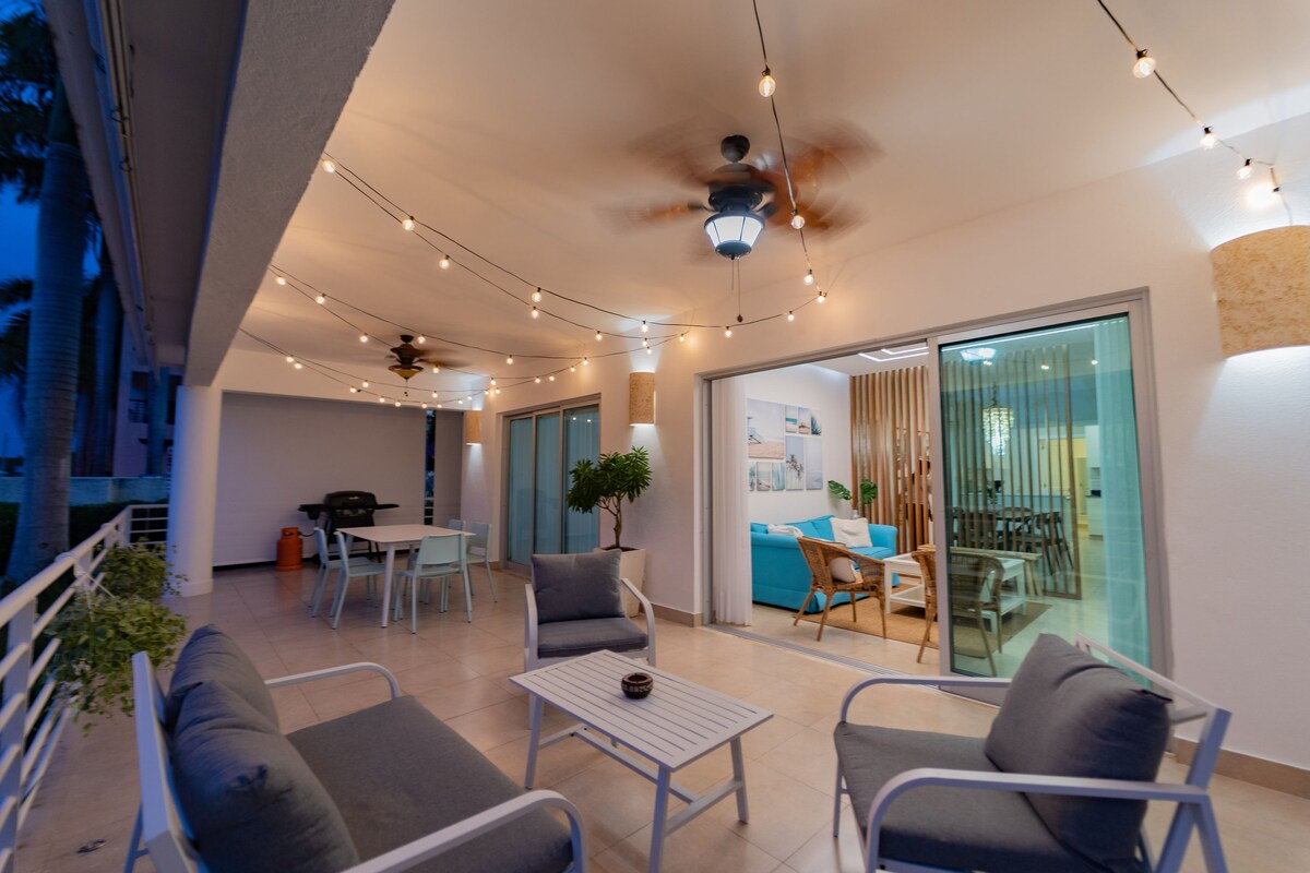 An outdoor patio is illuminated by string lights, creating a peaceful ambiance. Comfortable seating is arranged around a small table, with a barbecue grill visible in the background. Large sliding glass doors lead into the interior space, enhancing the connection between indoor and outdoor areas.