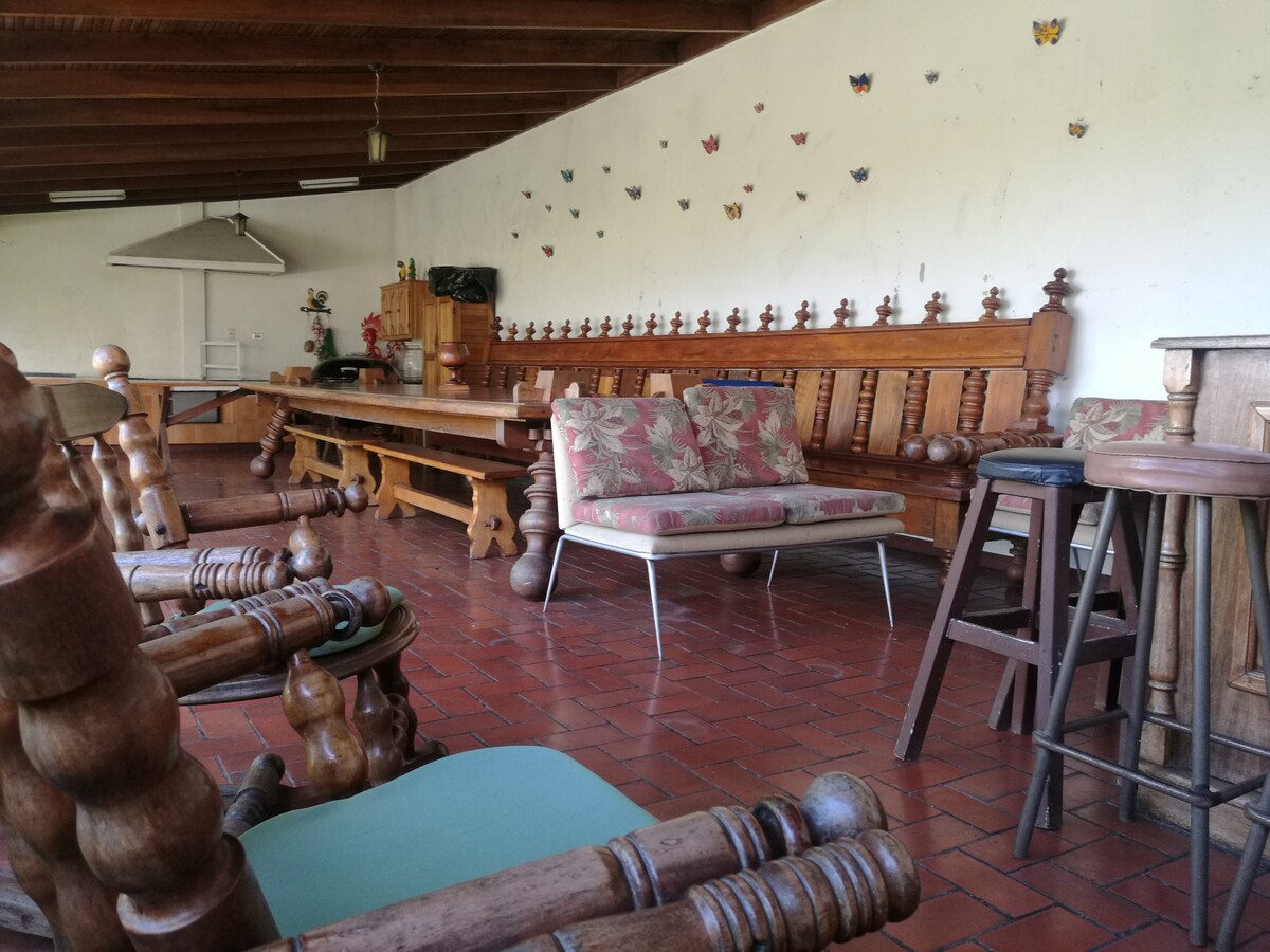 A spacious seating area features a long wooden table and multiple sturdy wooden chairs arranged around it. A comfortable sofa with a floral pattern is positioned nearby, with a collection of decorative butterflies adorning the white wall above.