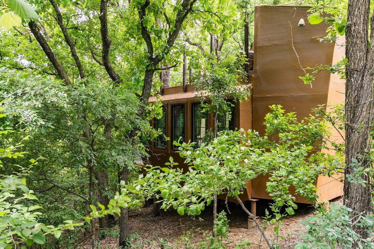 A treehouse blends harmoniously with the surrounding greenery, featuring earthy tones that complement the natural environment. Large windows provide views of the trees, and the structure is nestled among lush foliage, creating a private and serene atmosphere.