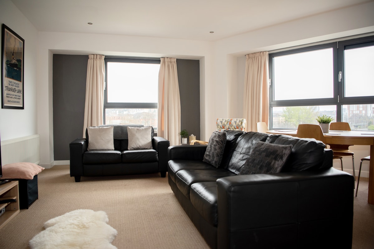 A modern living area features two black sofas positioned across from large windows adorned with light curtains. A small decorative plant and a patterned chair add a touch of warmth, while a dining table can be seen in the background.
