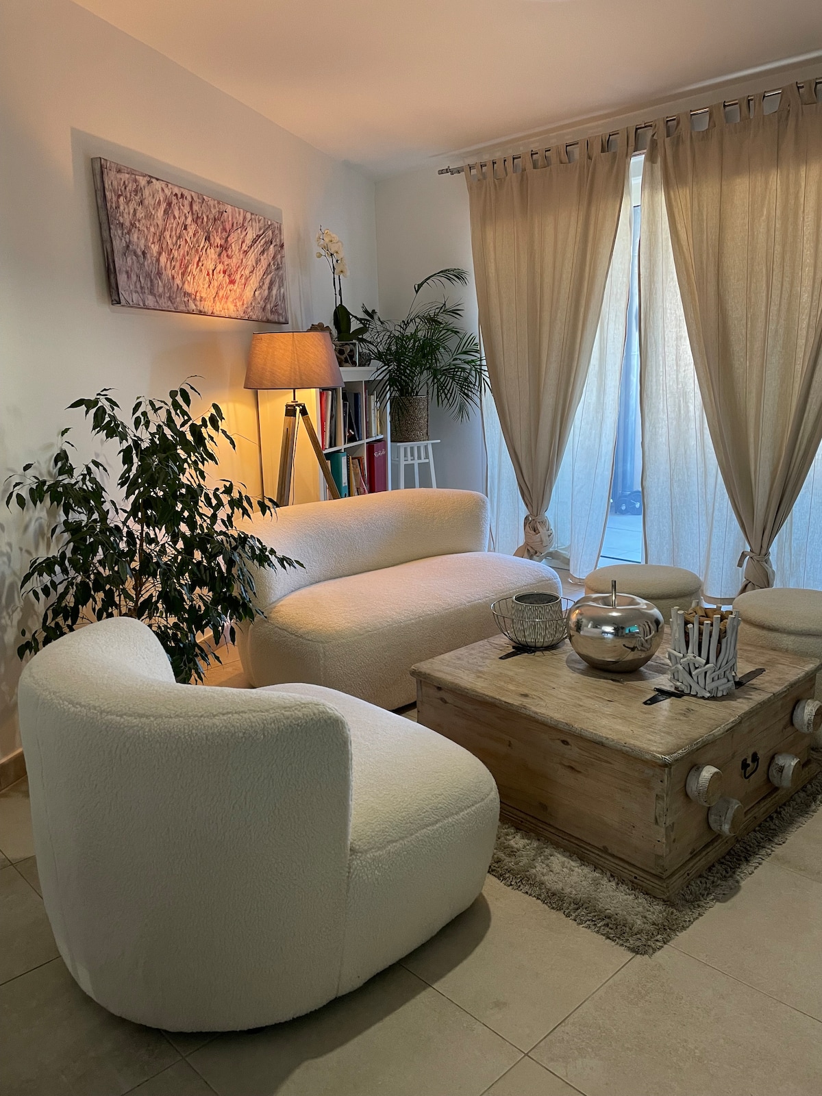 A seating area features two curved chairs and a cozy sofa, complemented by a wooden coffee table. Natural light filters through sheer curtains, illuminating a green plant and a decorative bowl. A lamp adds soft lighting, creating a relaxing environment.