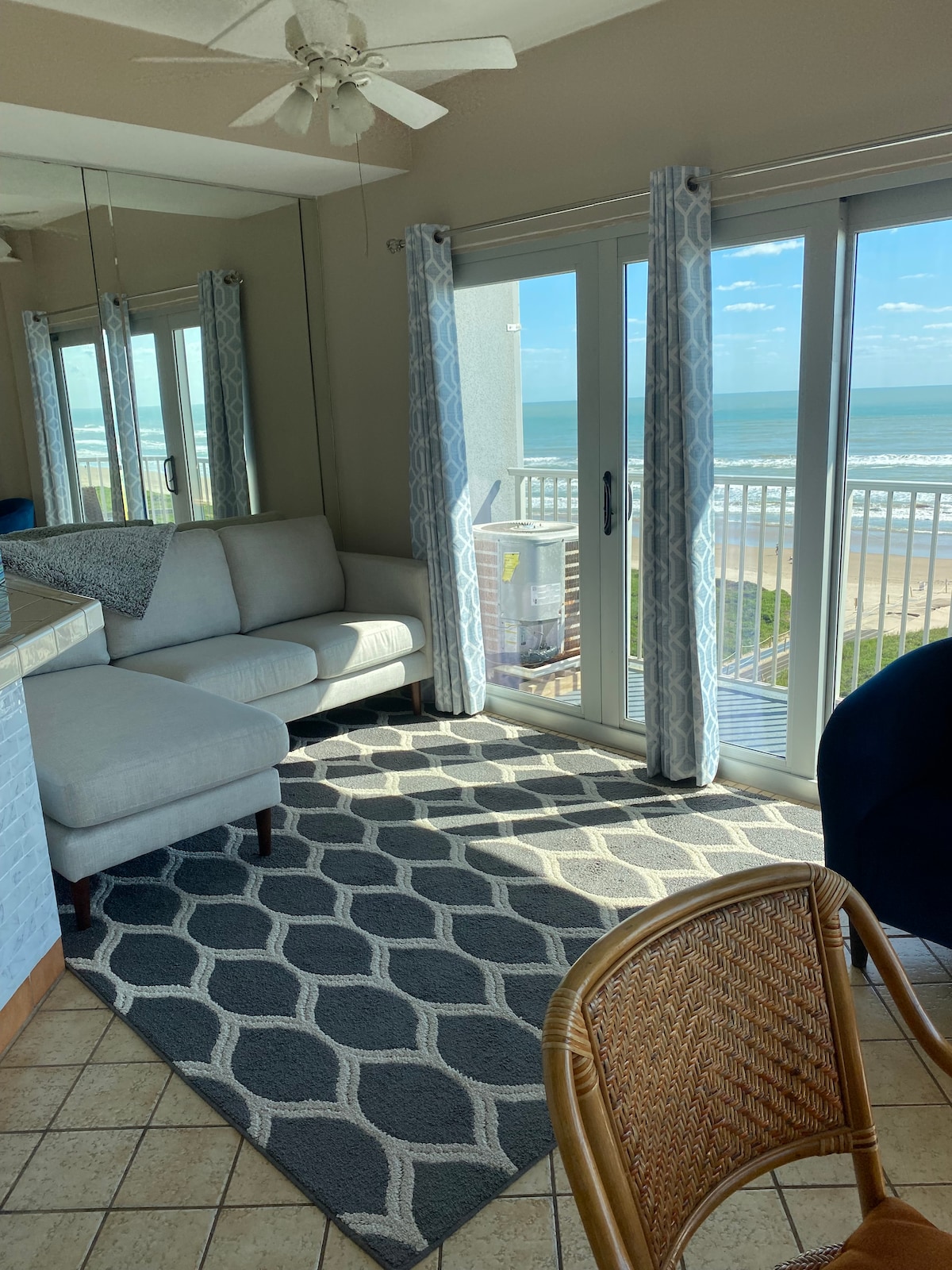 A bright living area features a light-colored sectional sofa facing large glass doors that open to a balcony with ocean views. Soft, patterned rugs cover the tiled floor, and sheer curtains frame the doors, allowing natural light to fill the space.