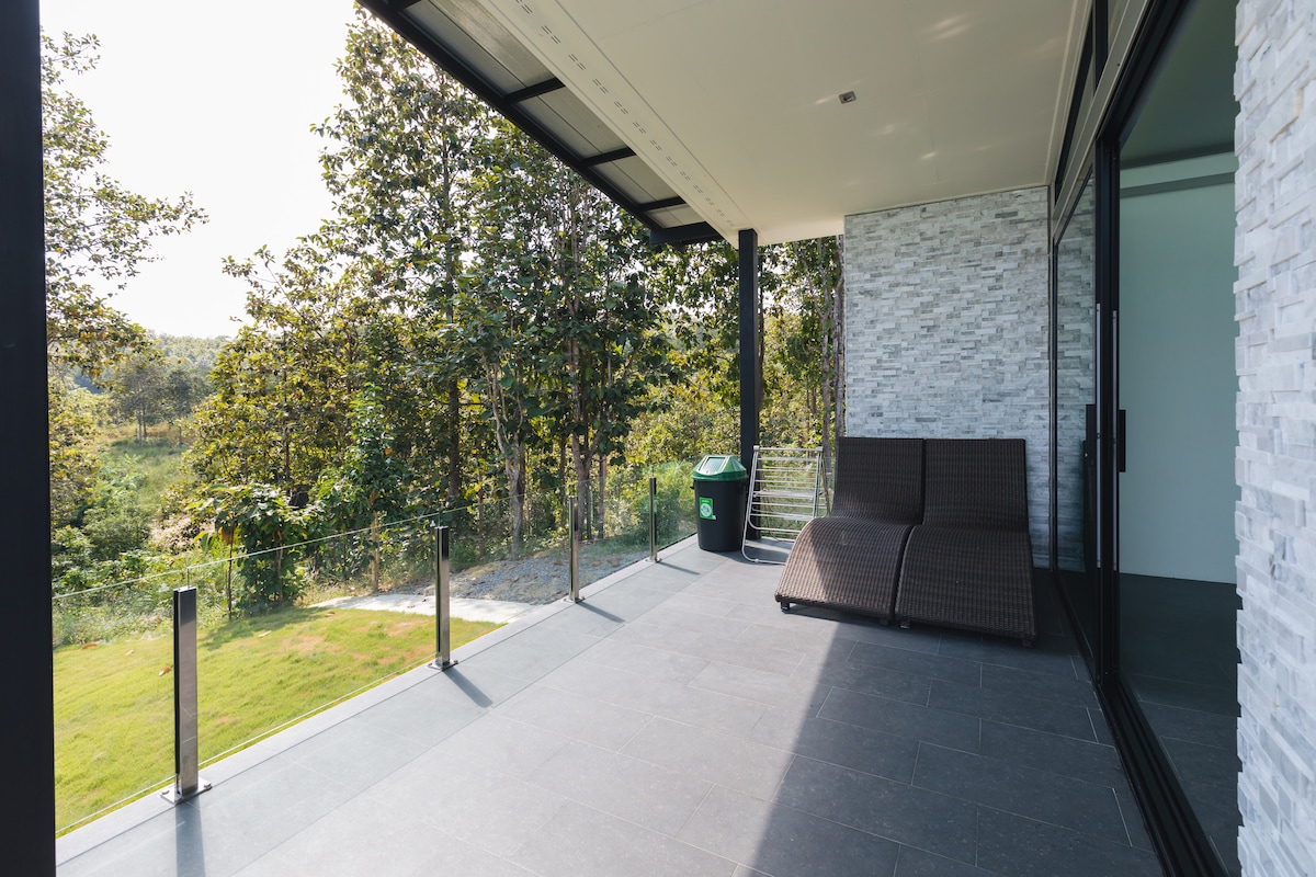A spacious terrace offers a comfortable seating area with a modern lounge chair, surrounded by lush greenery. Large sliding glass doors provide access to the interior, emphasizing a seamless transition between indoor and outdoor living.