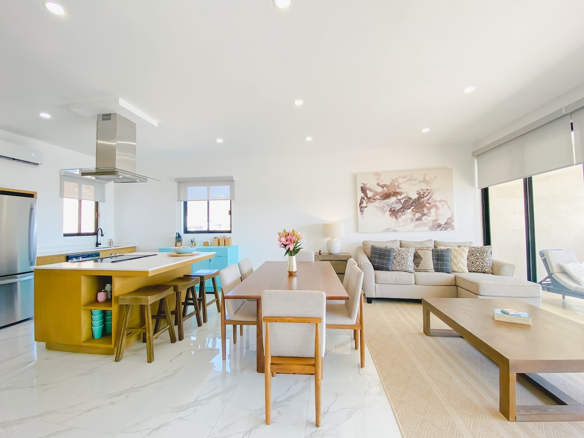 A modern open living space incorporates a kitchen with a breakfast bar and seating for six. A spacious seating area features a sectional sofa adorned with textured cushions. Natural light fills the room through large windows, enhancing the contemporary design.