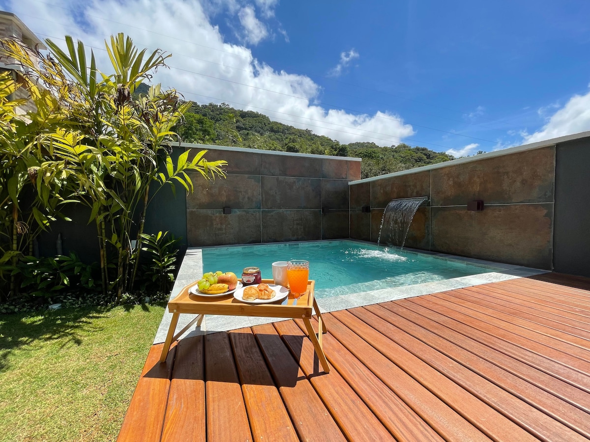 A private pool is framed by wooden decking and lush greenery, featuring a waterfall feature. A stylish tray with breakfast items and beverages is set nearby, inviting relaxation in a sunlit environment under a clear blue sky.