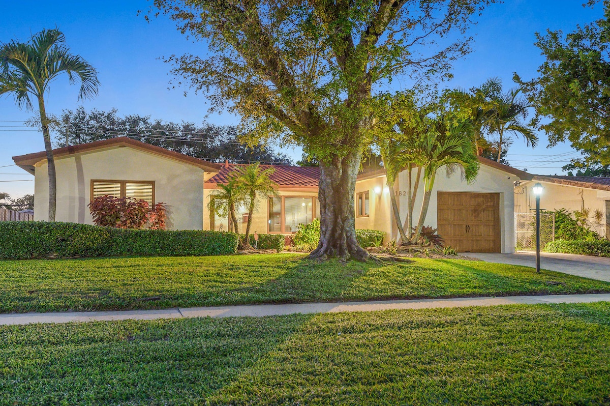 The exterior of a modern home is framed by lush greenery and palm trees. A spacious lawn leads to the entrance, where a welcoming front porch can be seen. The warm glow of outdoor lighting highlights the home's features as evening sets in.