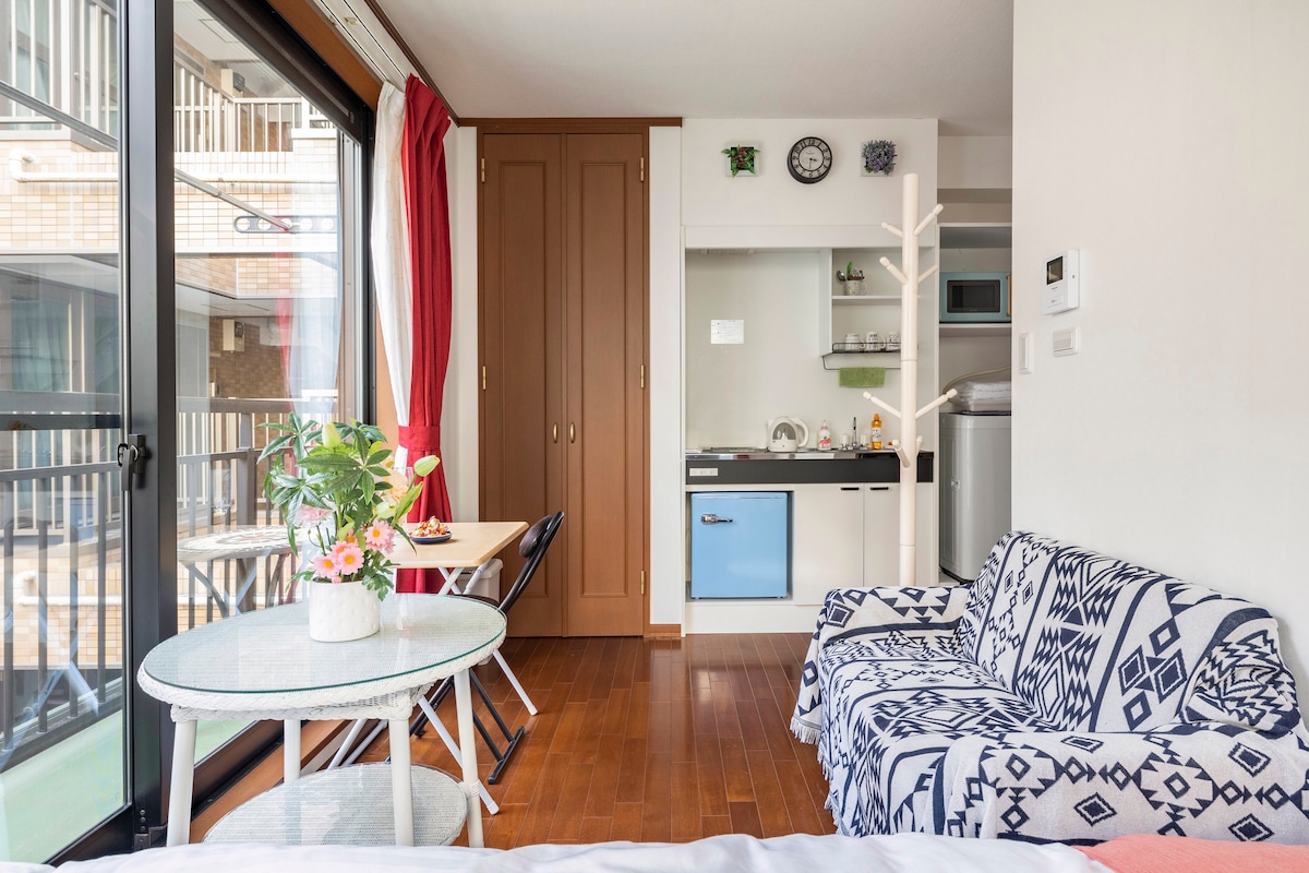 An inviting living area features a cozy sofa draped with a patterned throw, complemented by a small dining table arranged with a potted plant. The kitchenette is visible in the background, showcasing modern appliances against light cabinetry. Large windows allow ample natural light.
