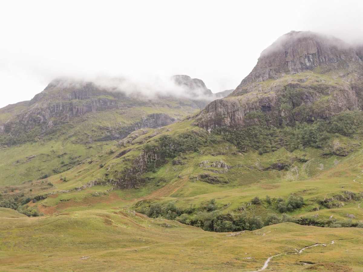 Rolling hills covered in lush greenery are framed by towering mountains draped in mist. A winding path can be seen meandering through the landscape, inviting exploration. The natural beauty of the area reflects the serene atmosphere typical of Glencoe.