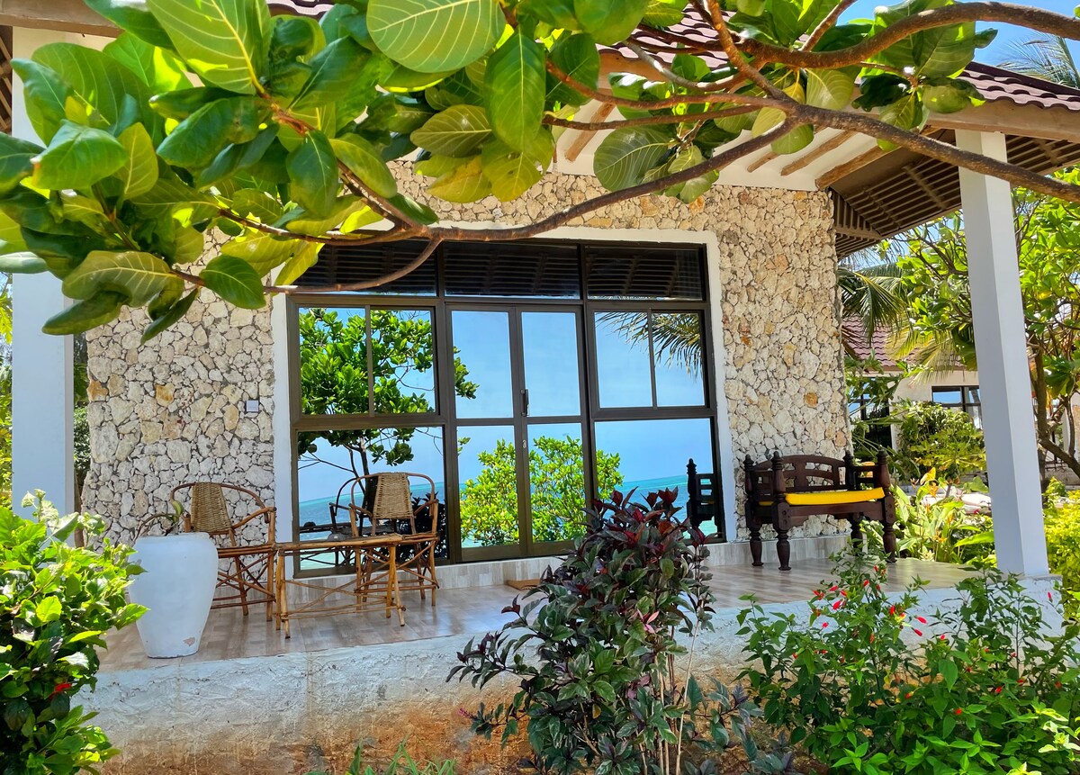 A charming outdoor seating area is framed by lush greenery and vibrant flowers. The view features large glass windows, offering a glimpse of the ocean in the distance. Natural stone walls complement the wooden furniture, creating a serene atmosphere near the water.