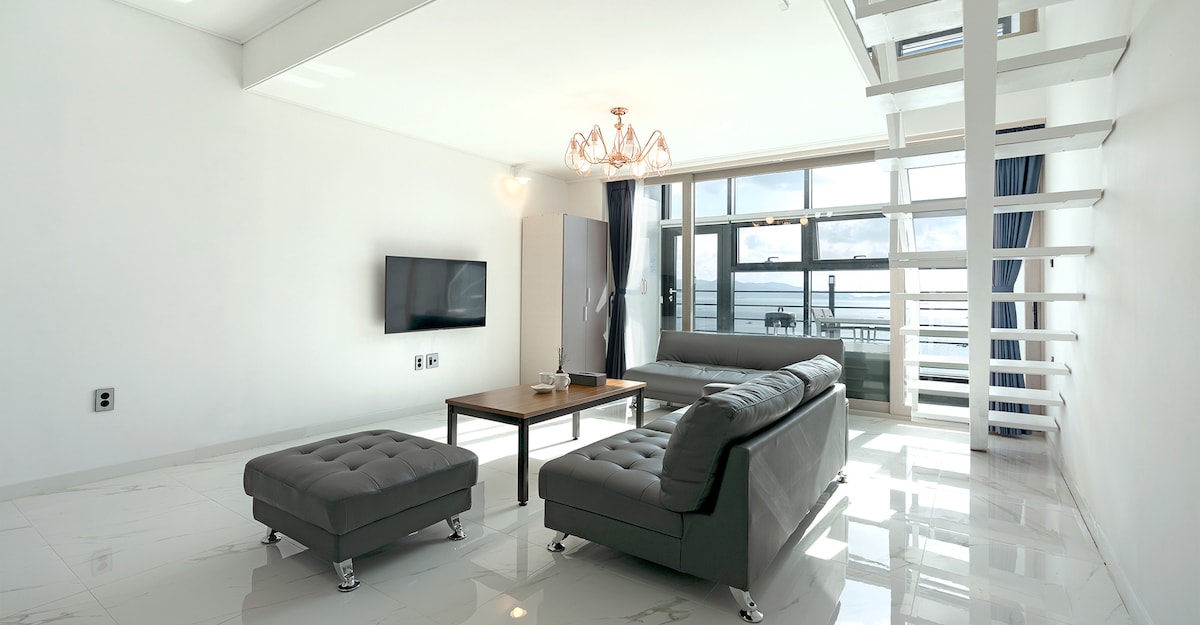 A spacious living area features large windows that provide views of the ocean. A two-tone gray sofa is positioned around a wooden coffee table. A sleek television is mounted on the wall, with a modern chandelier hanging above. A staircase leads to the upper level.