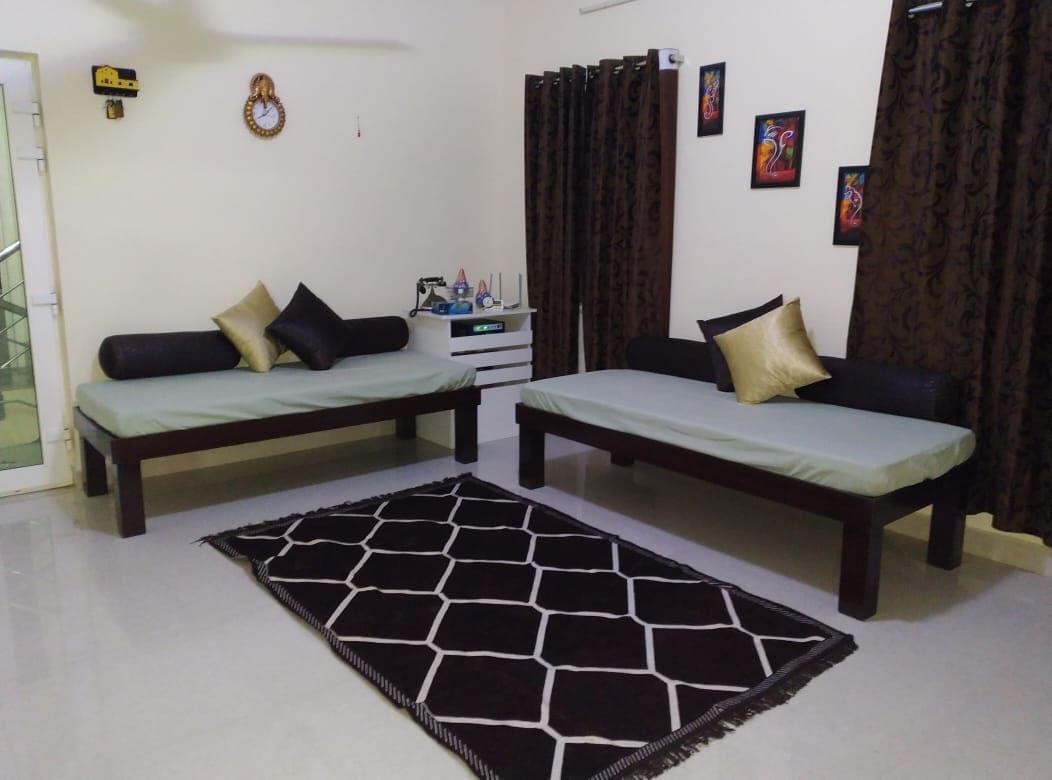 A light-filled living area is furnished with two daybeds, each adorned with decorative pillows. A dark rug with a geometric pattern anchors the space, while two pieces of vibrant wall art add a touch of color. Nearby, curtains in a rich hue frame the windows.