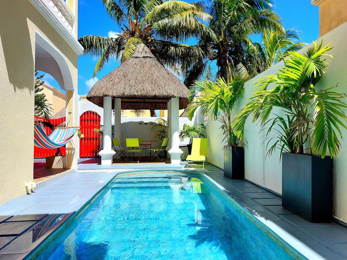 A tranquil pool area is surrounded by lush palm trees and well-maintained plants. A thatched gazebo is featured alongside the pool, complemented by bright seating. Sunlight reflects off the water, creating a peaceful atmosphere perfect for relaxation.