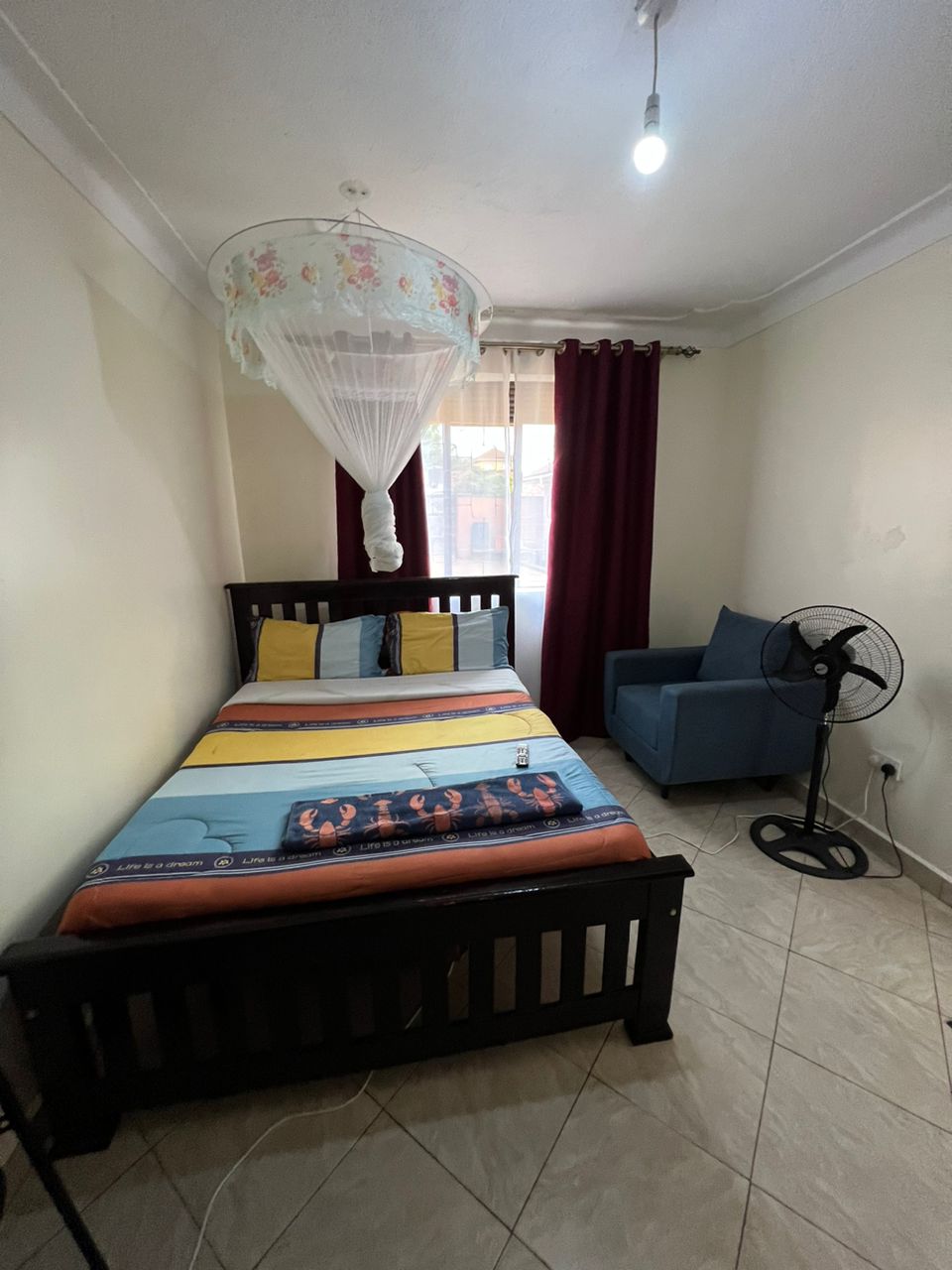 A studio bedroom features a compact bed adorned with patterned bedding, a decorative canopy above. A blue armchair is positioned near a window covered by light curtains. A standing fan and dark red curtains contrast with the neutral walls, creating a simple yet functional space.
